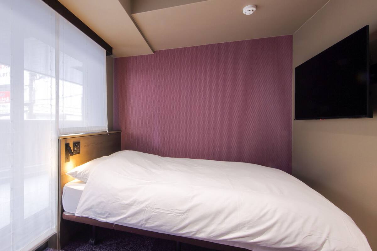 A small, private room with a bed, a window with blinds, a wall-mounted TV, and a ceiling light.