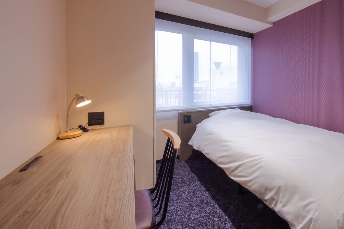 A hotel room with a bed, a desk, a chair, a window, and a purple wall.
