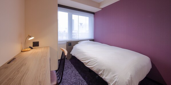 A hotel room with a bed, a desk, a chair, a window, and a purple wall.