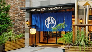The exterior of Hotel Sui Kobe Sannomiya with a blue banner displaying the hotel's logo, a wooden planter with plants, and a well-lit entrance. - HOTEL SUI KOBE SANNOMIYA by ABEST (Kobe)