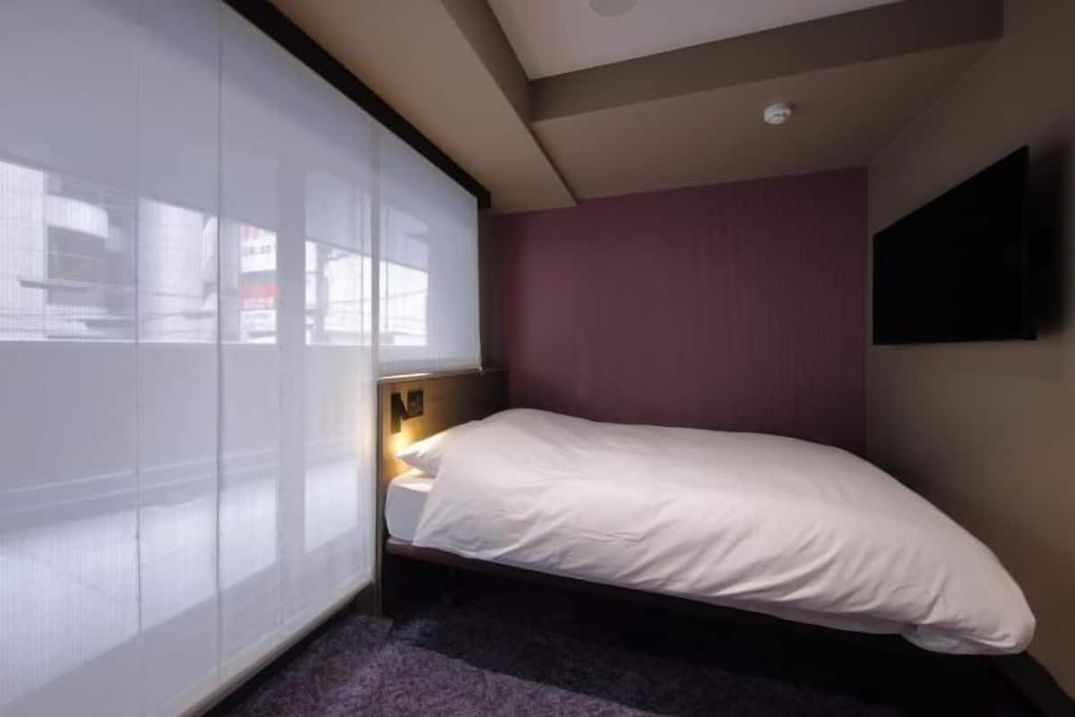 A bedroom with a bed, a nightstand, a window with blinds, and a wall-mounted TV.