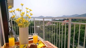 A balcony with a wooden table set with breakfast items, a vase of yellow flowers, and a scenic view of mountains and greenery. - Apartment Kola  (Dubrovacko Primorje)
