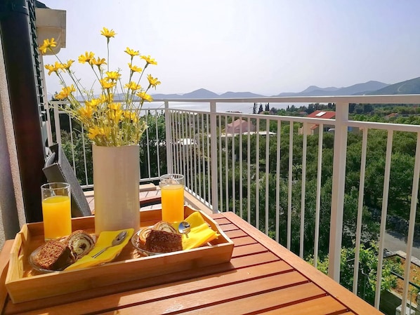 A balcony with a wooden table set with breakfast items, a vase of yellow flowers, and a scenic view of mountains and greenery. - Apartment Kola (Dubrovacko Primorje)