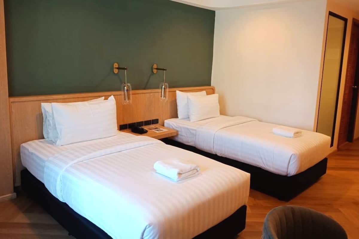 Habitación de hotel con dos camas individuales, cabecera de madera, pared de acento verde y una mesita de noche con lámpara.
