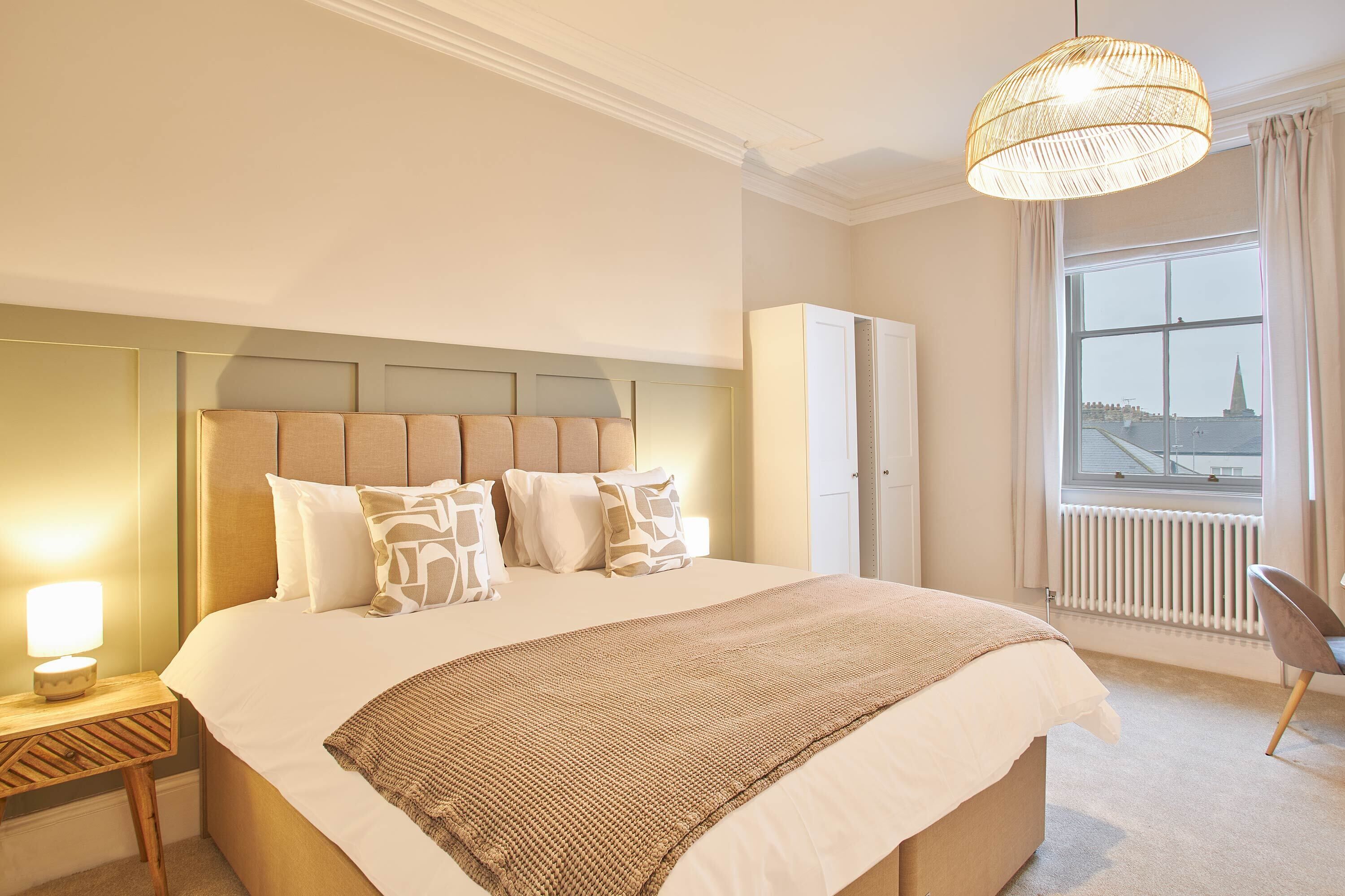Een keurig opgemaakt slaapkamer met een groot bed, een bedlampje en een uitzicht op een stadsgezicht door een raam.
