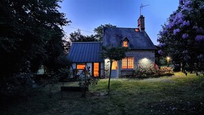 Een stenen huis met een schuin dak en schoorsteen, omgeven door een tuin met bloeiende planten en een bank.
