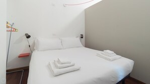 Een net opgemaakt bed met witte lakens en gevouwen handdoeken, een bedlamp en een rode plank.