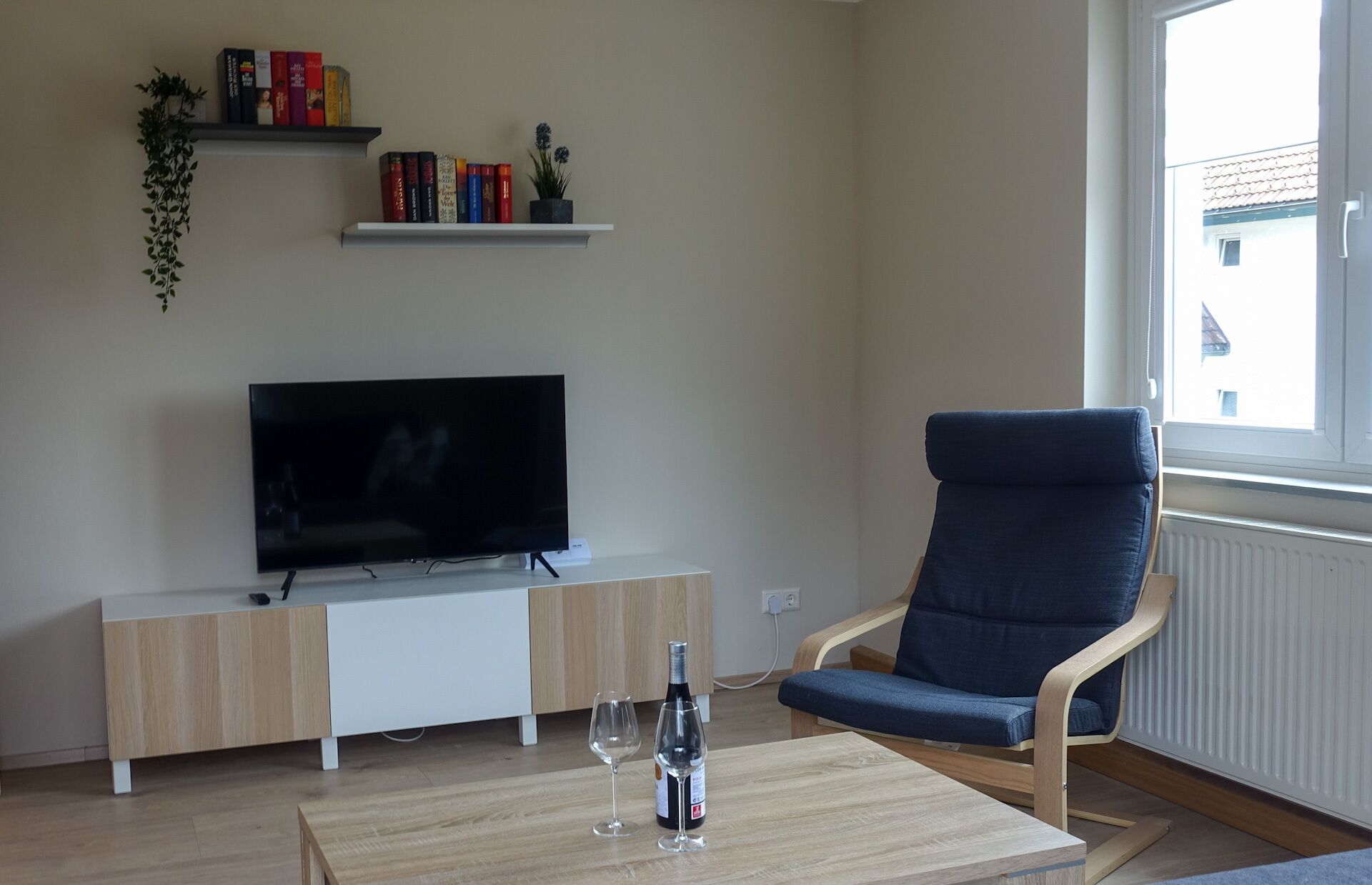 A living room with a flat-screen TV on a white cabinet, a blue armchair, a wooden coffee table with a wine bottle and glass, and a window with a view of a building outside.