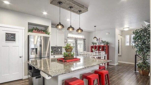 Uma cozinha moderna com bancada de granito, eletrodomésticos de aço inoxidável, banquetas vermelhas, iluminação pendente e um vaso de planta.