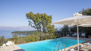 Una piscina azul cristalina con baldosas de piedra, rodeada de sillones de descanso y sombrillas, con vistas a un paisaje montañoso.