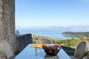 A table with a bowl of fruit, a bottle, and a glass, overlooking a scenic view of a body of water and hills. - Villa Pietra Gialla by Rodostamo Collection (Corfu)