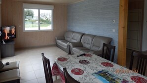 A living room with a sofa, a TV, a dining table with chairs, and a window with a view of greenery. - FREHEL HOUSE (Fréhel)