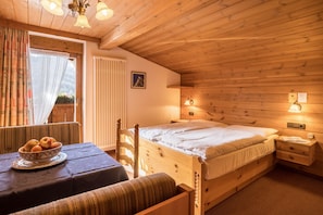 A wooden room with a bed, a table with a bowl of fruit, and a window with curtains.