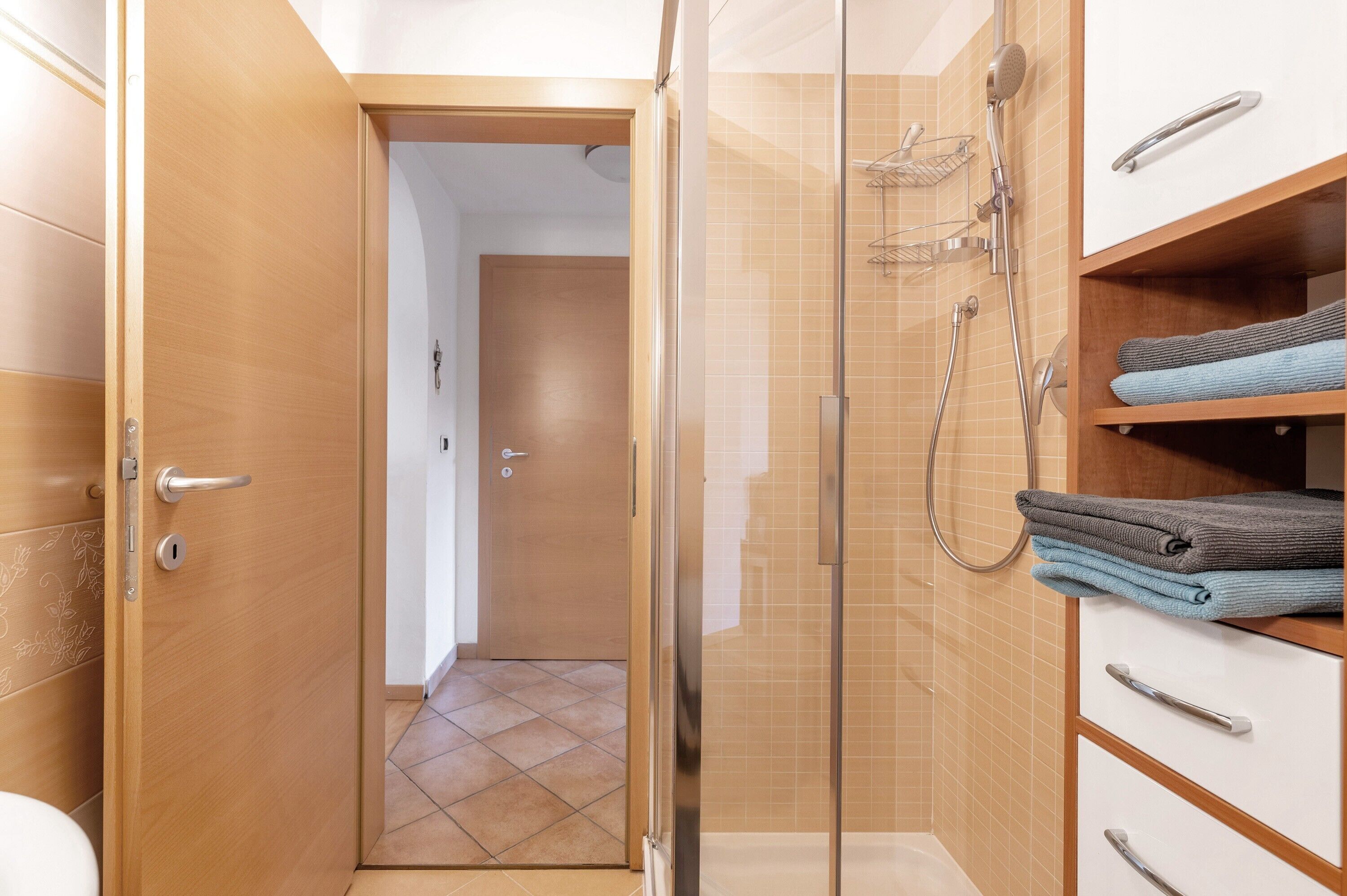 Een badkamer met een douche in een glazen cabine, een houten deur, een betegelde vloer en een handdoekrek met opgevouwen handdoeken.