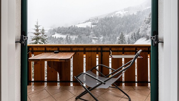 Ein Balkon mit Holztisch und Metallstuhl, der auf eine verschneite Landschaft blickt.