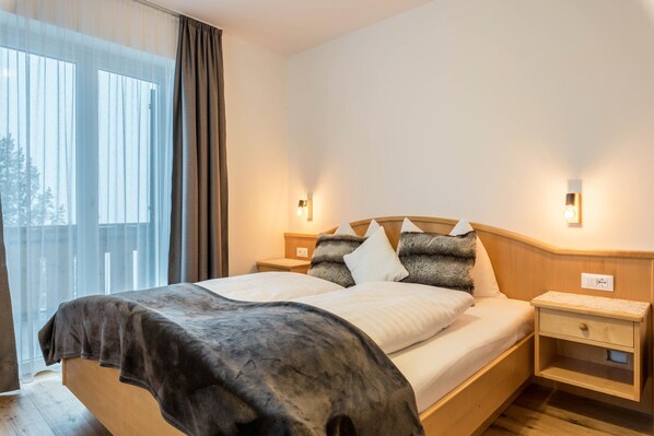 A bedroom with a bed, two bedside tables, a nightstand, a window with curtains, and a view of trees outside.
