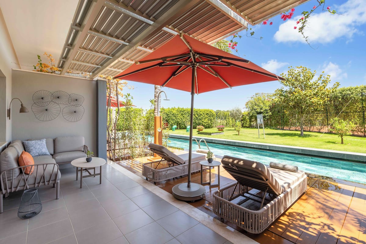 Um pátio moderno ao ar livre com piscina, incluindo um sofá, espreguiçadeiras e um guarda-sol vermelho.
