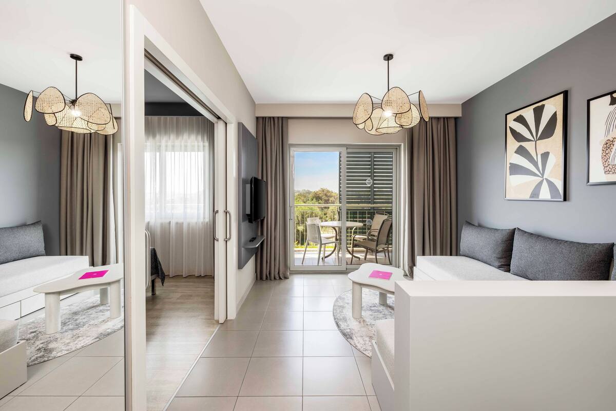 Quarto de hotel moderno com sofá, televisão, área de refeições e varanda com cortinas.