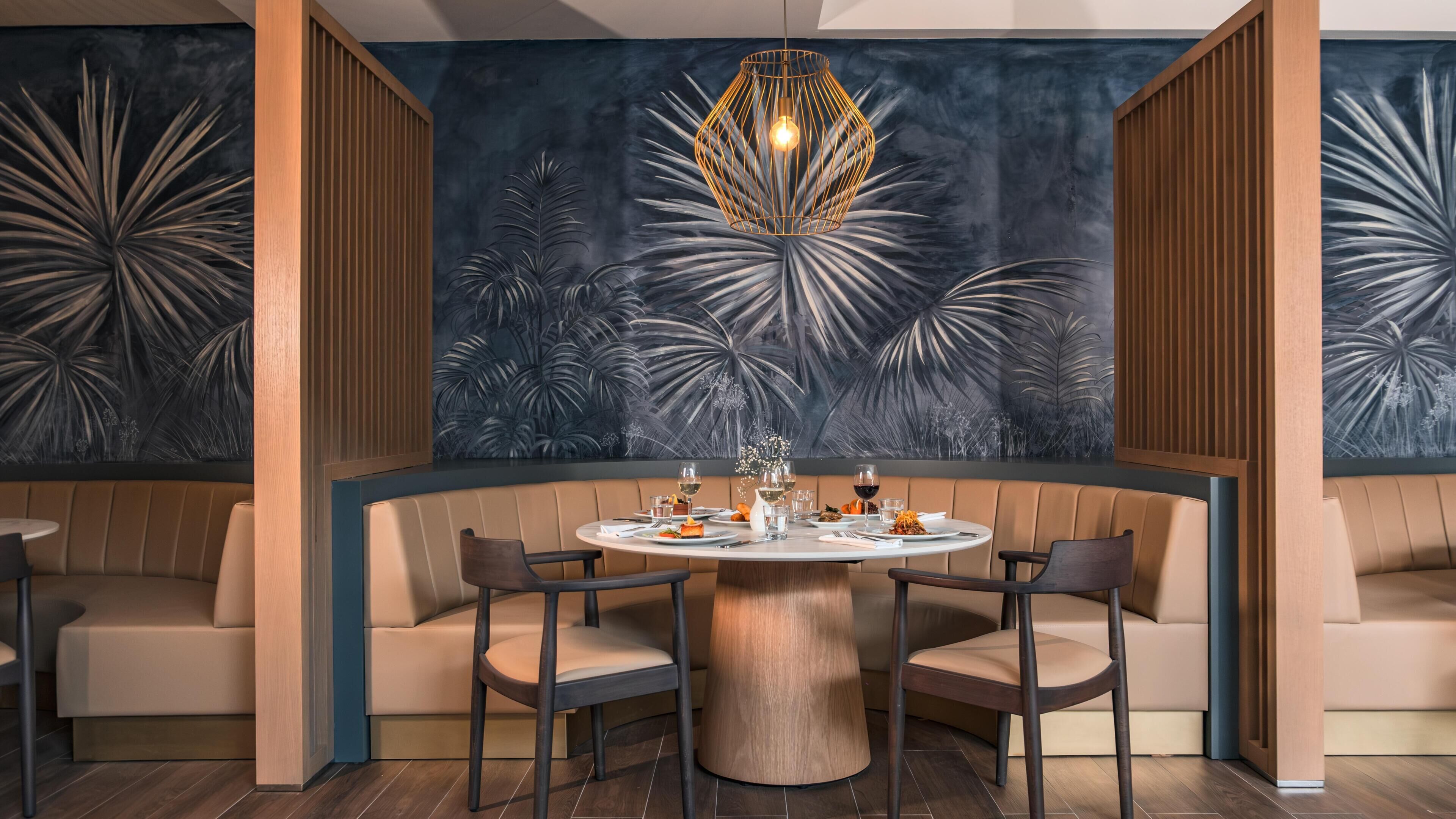 Una mesa redonda con seis sillas, una lámpara colgante en el centro y una pared con diseño de hojas tropicales.