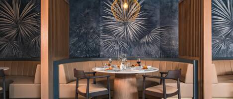 Una mesa redonda con seis sillas, una lámpara colgante en el centro y una pared con diseño de hojas tropicales.