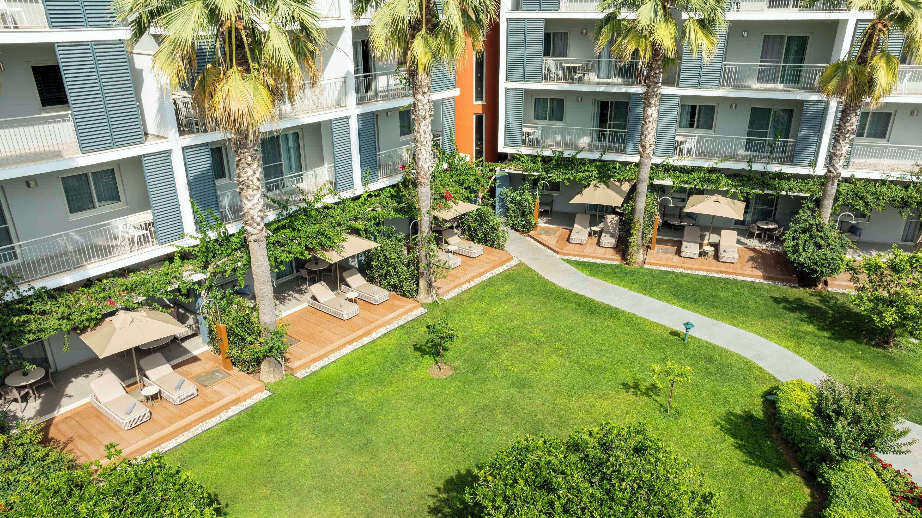 Un hôtel à plusieurs étages doté d’espaces extérieurs aménagés pour se détendre, de palmiers et d’une pelouse bien entretenue.