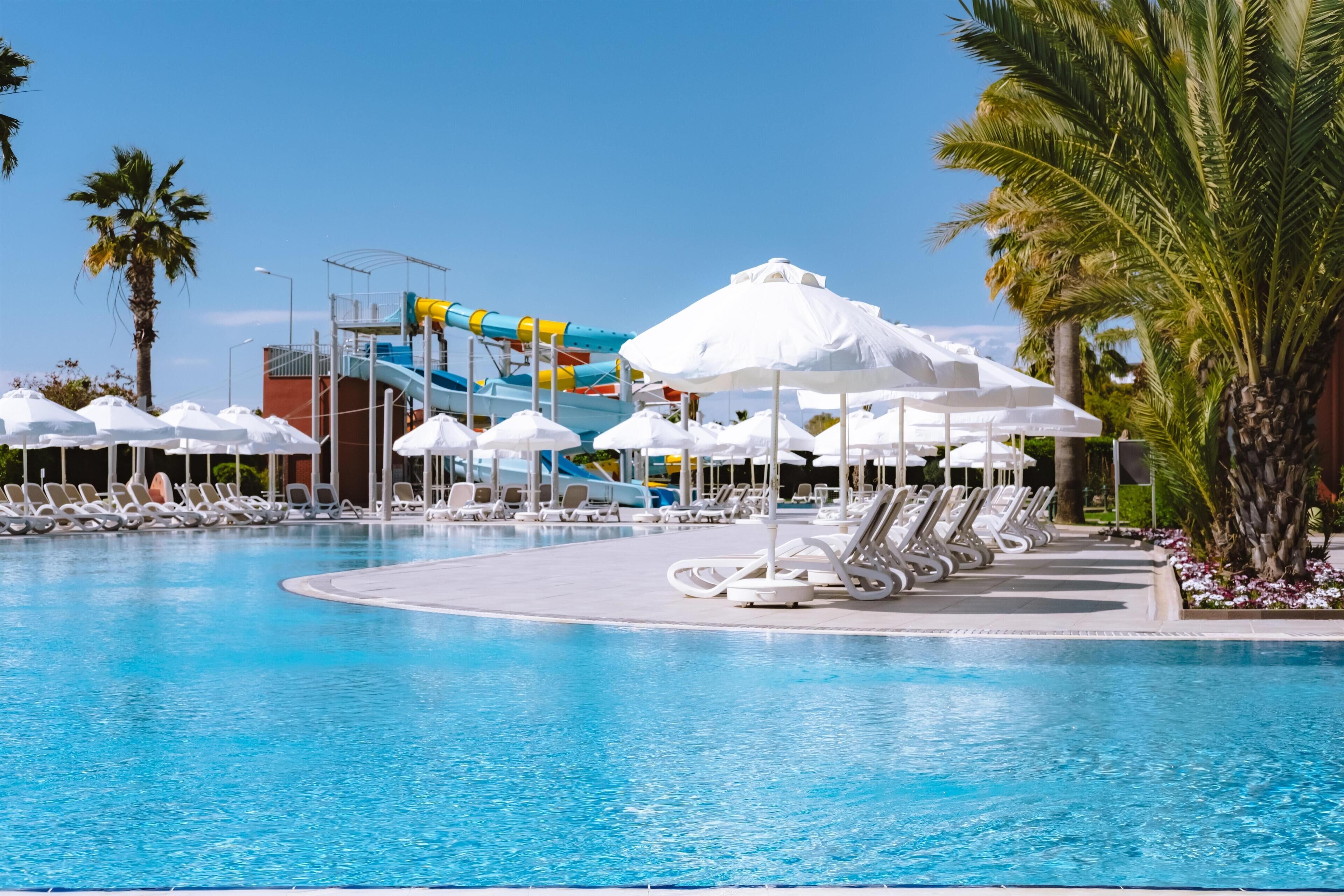 A pool area with a water slide, lounge chairs, umbrellas, and palm trees.