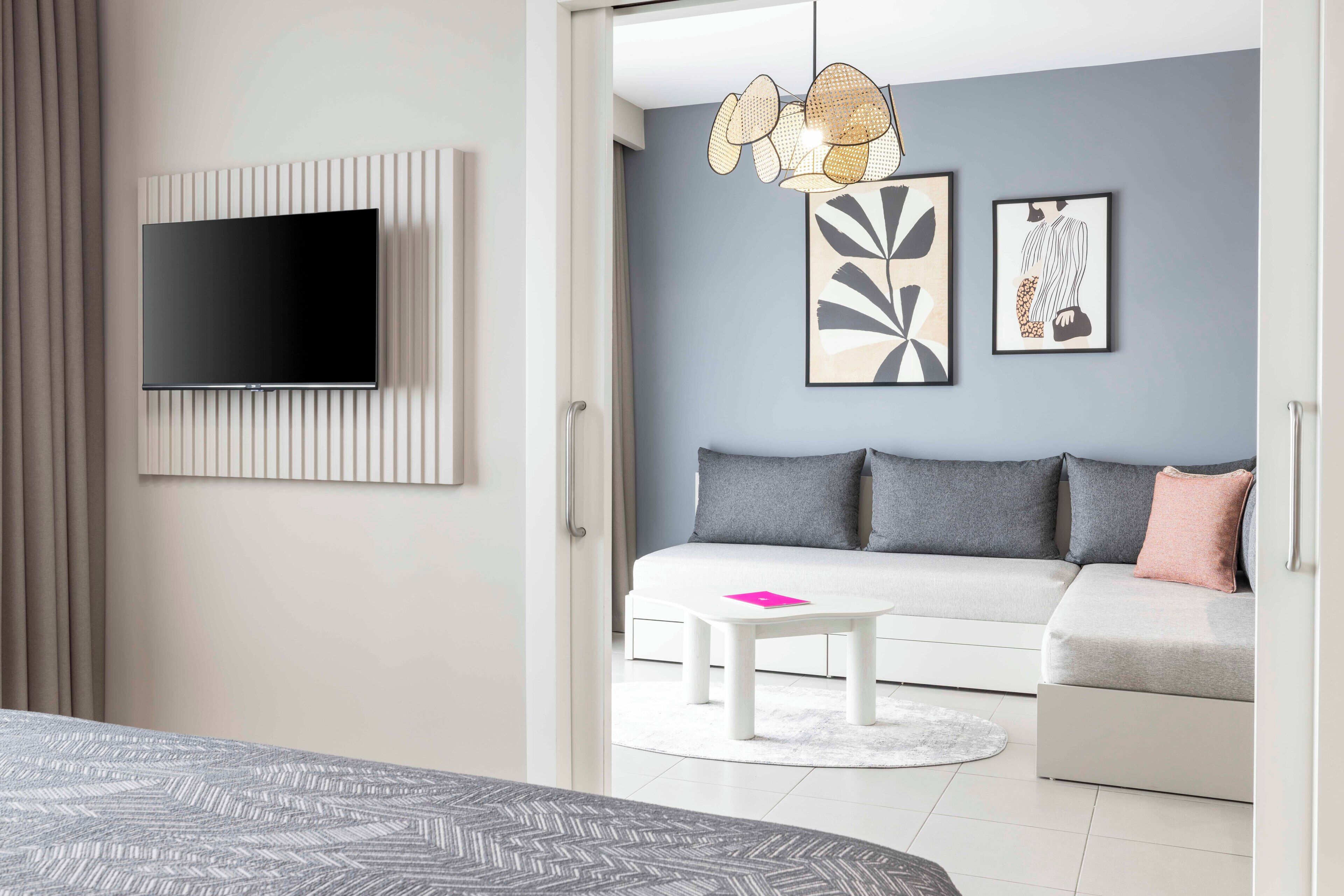 A modern hotel room with a flat-screen TV mounted on the wall, a sofa, a small white table, and a decorative hanging light fixture.