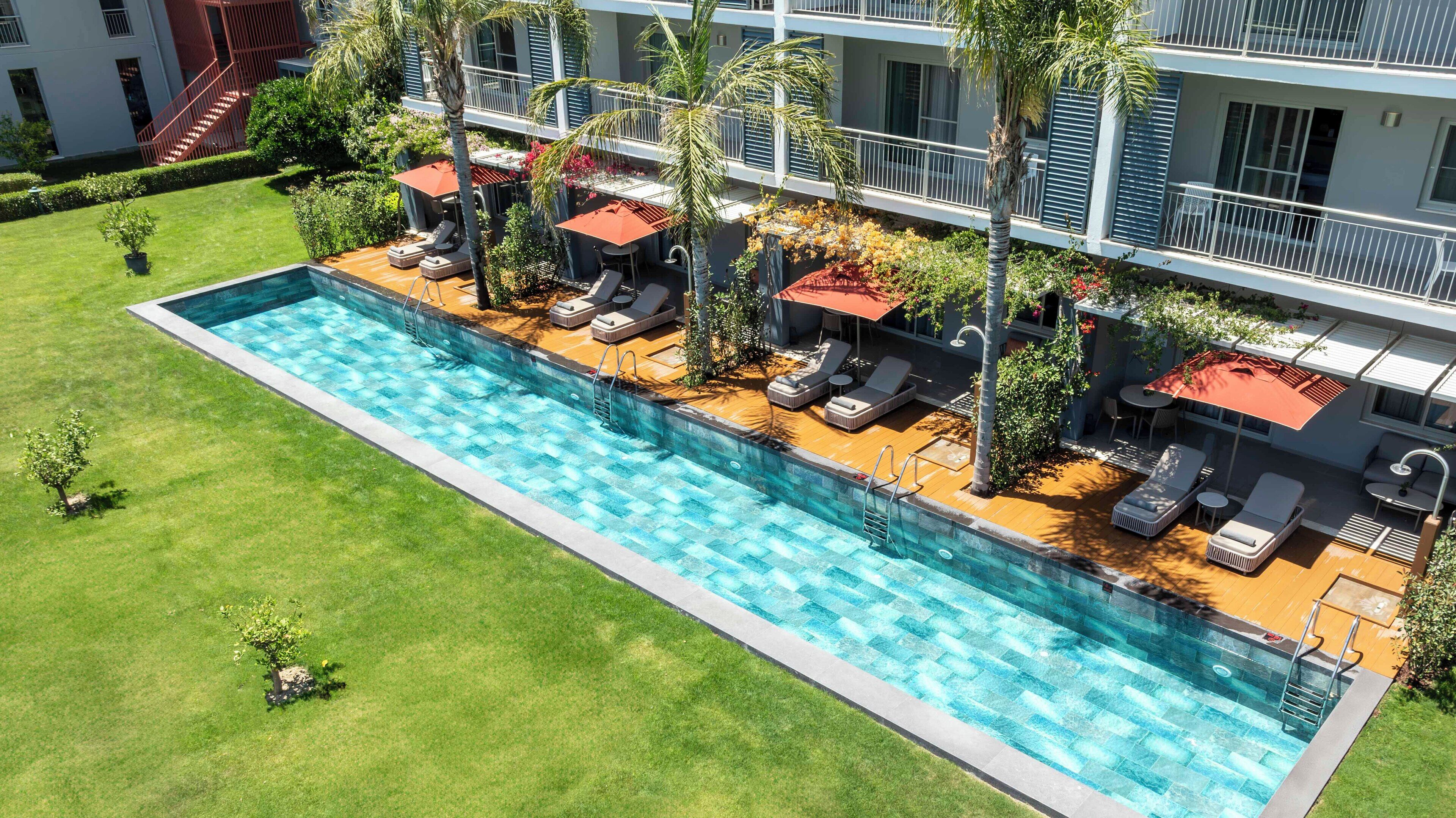 Une piscine entourée de chaises longues et de parasols, elle-même bordée d’une pelouse bien entretenue et d’un immeuble à plusieurs étages doté de balcons.