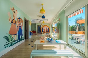 A brightly lit children's playroom with a mural, tables, benches, and large windows.