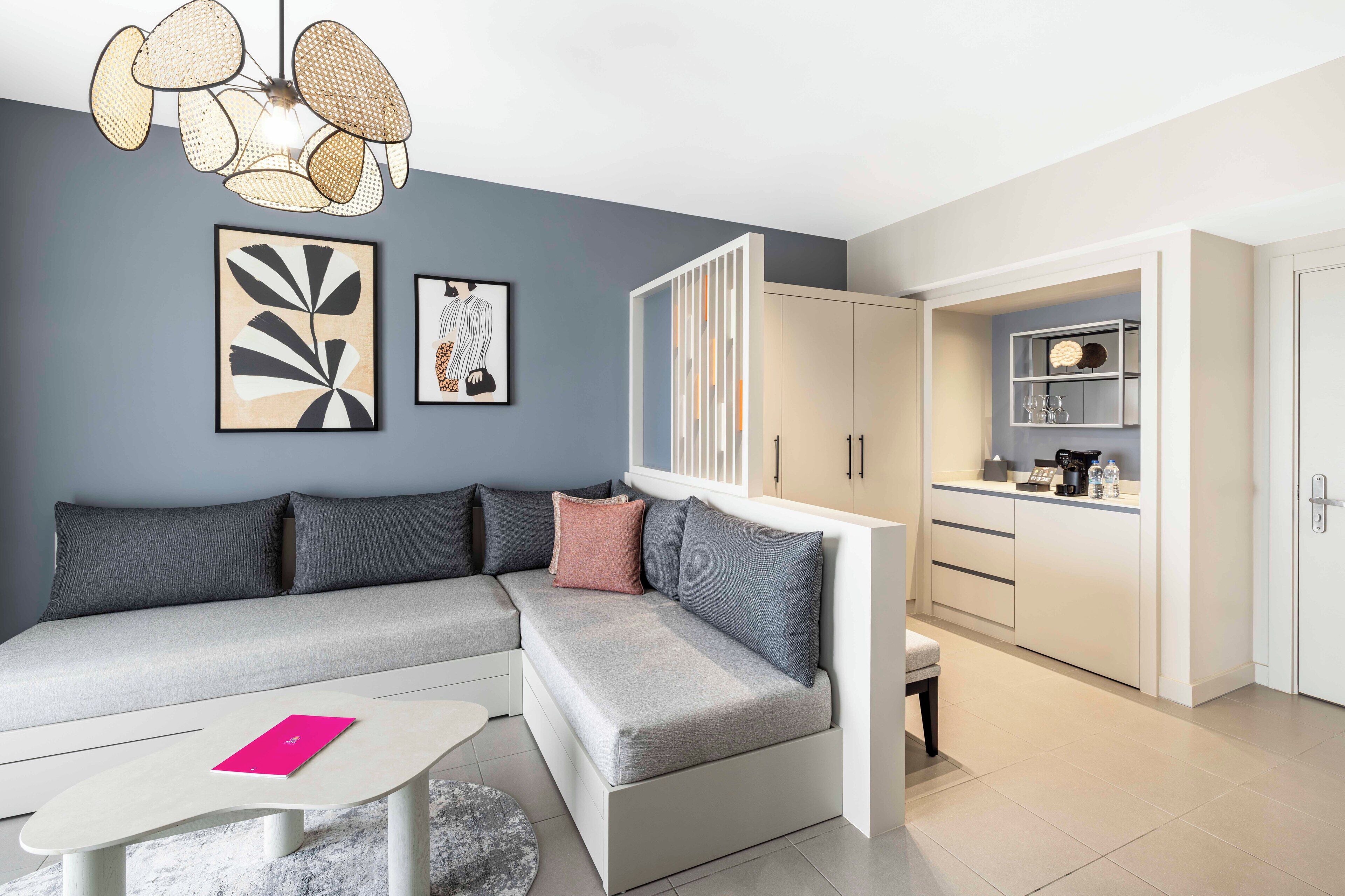 A modern living room with a grey sectional sofa, a white coffee table, and a kitchen area with white cabinets and a countertop.