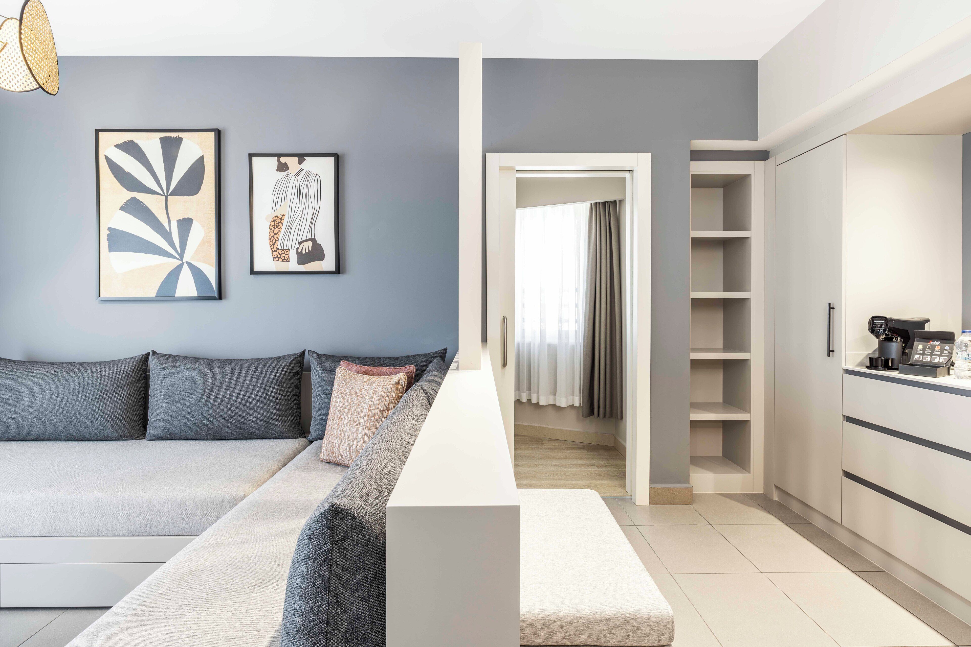 A modern living room with a sofa, a coffee table, and artwork on the wall. There is a kitchen area with cabinets and a countertop.