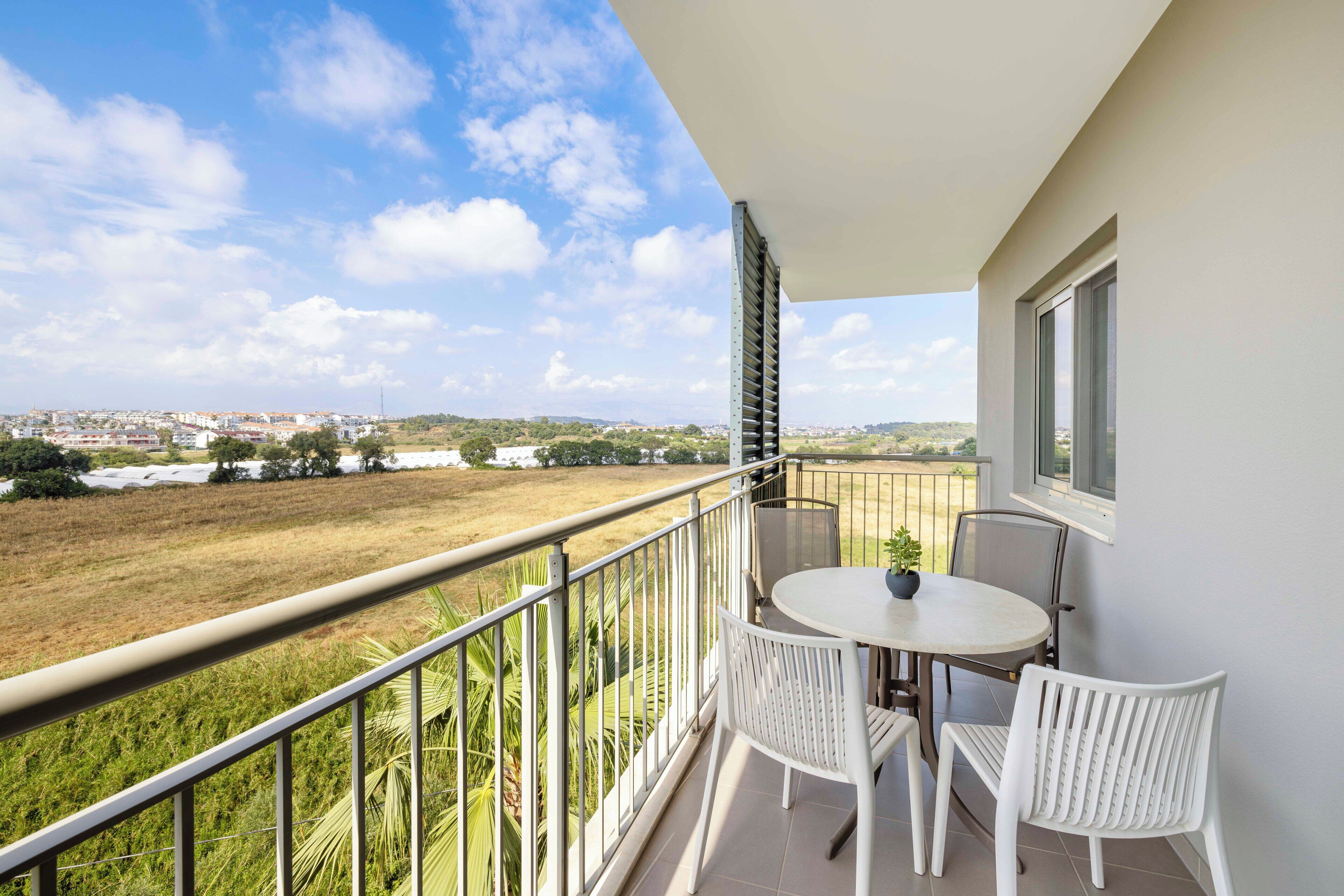 A balcony with a table and chairs, overlooking a grassy field and distant buildings.