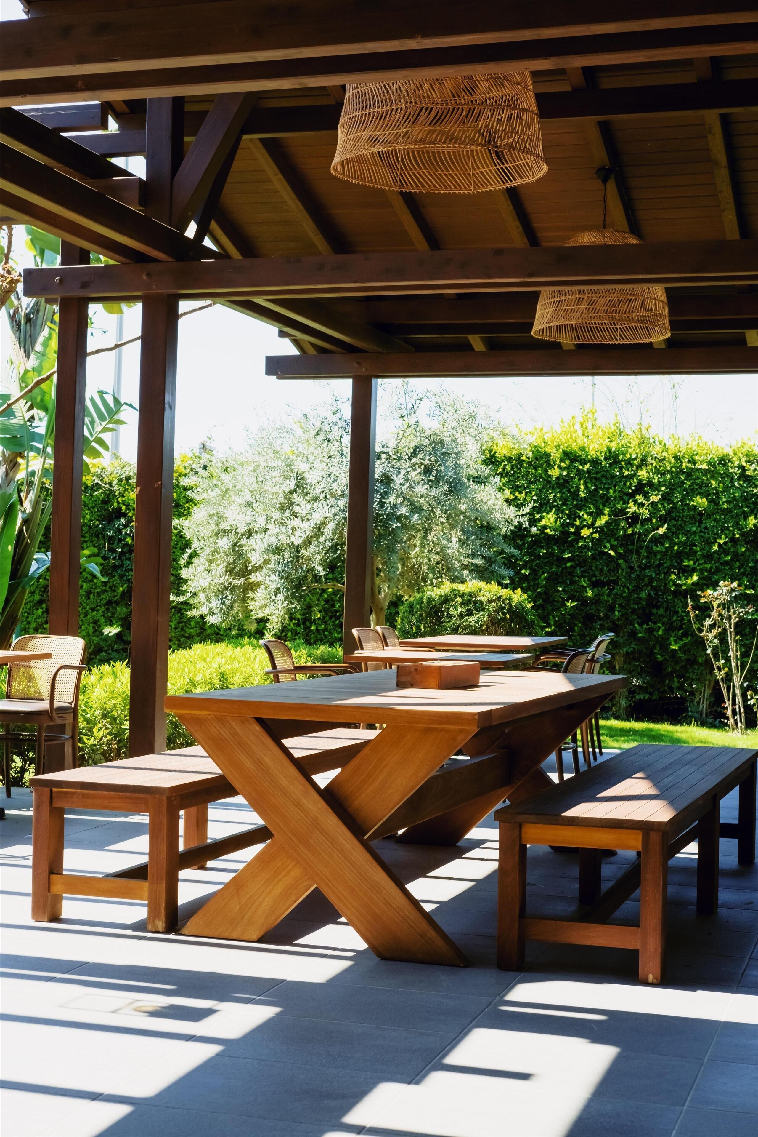 Un patio al aire libre con mesas y sillas de madera, una pérgola con lámparas de fibra y abundante follaje al fondo.