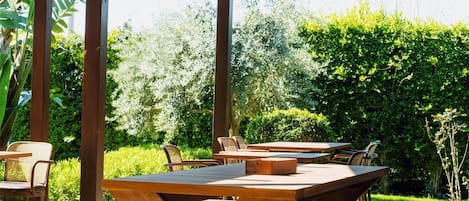 Un patio al aire libre con mesas y sillas de madera, una pérgola con lámparas de fibra y abundante follaje al fondo.