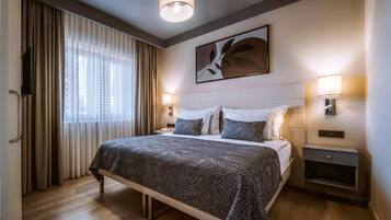 Una habitación de hotel moderna con una cama grande, dos mesitas de noche, un televisor montado en la pared y una ventana con cortinas.