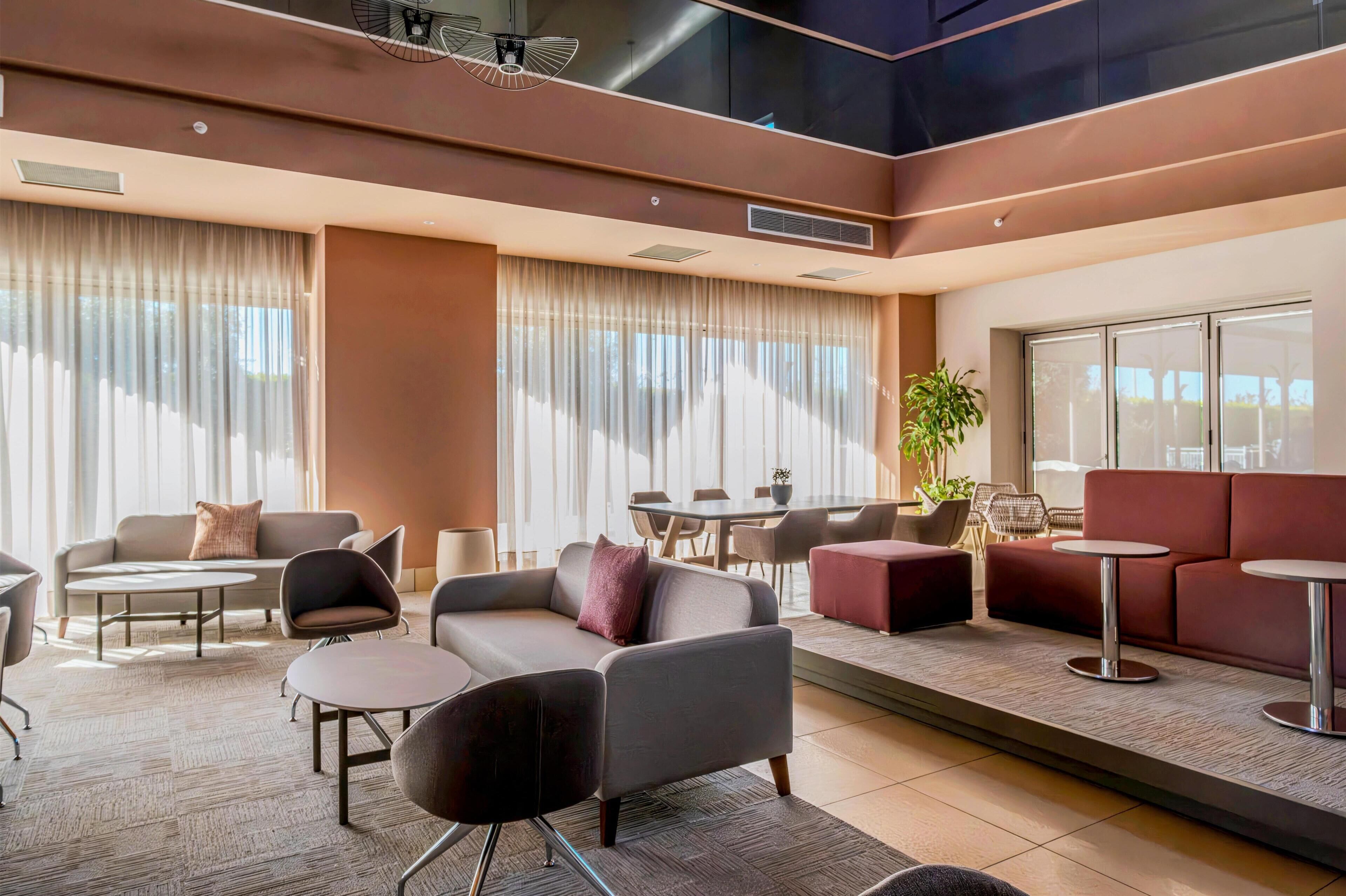 A modern lounge area with sofas, chairs, and tables, large windows with curtains, and a potted plant.