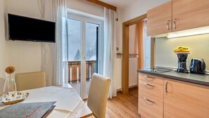 Eine moderne Küche mit Holzsärchen, einem Esstisch mit Stühlen, einem an der Wand montierten Flachbildfernseher und einem Balkon mit Blick auf Bäume.