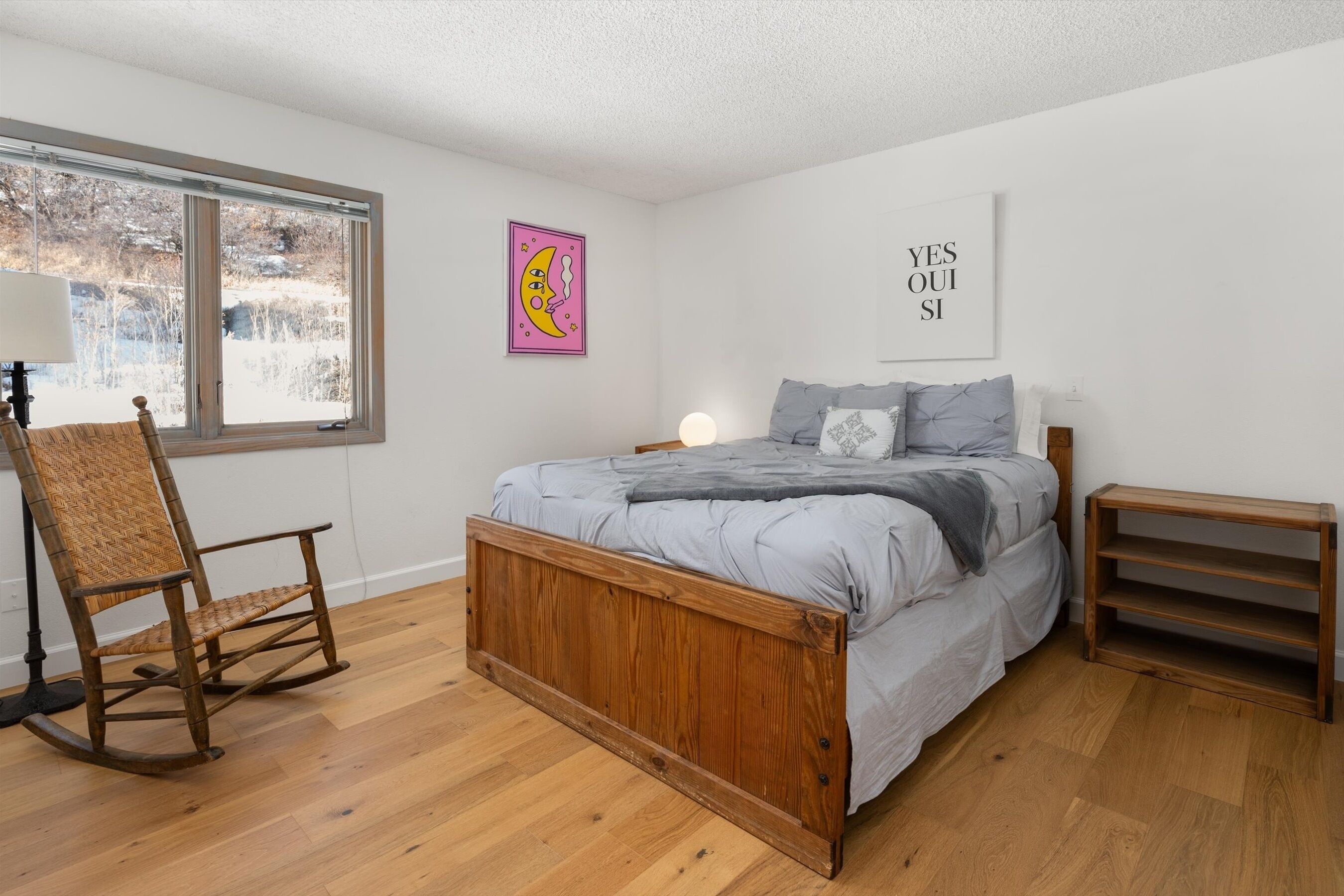 A bedroom with a wooden bed, a rocking chair, a bedside table, and a window with a view of trees.