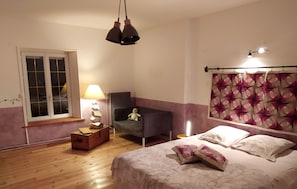 A bedroom with a wooden floor, a bed with patterned bedding, a chair with a teddy bear, a lamp, and a window with a view. - Chez Céline en campagne (Mazerny)