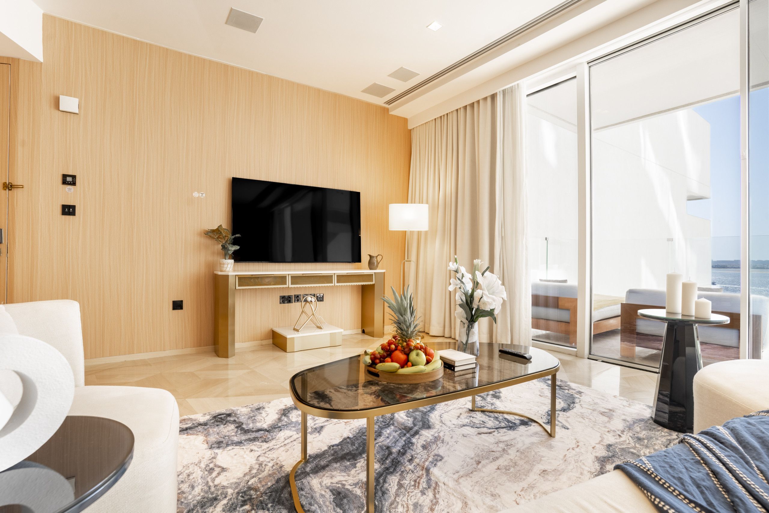 A modern living room with a flat-screen TV, a coffee table with fruit, a sofa, and a view of the outdoors.