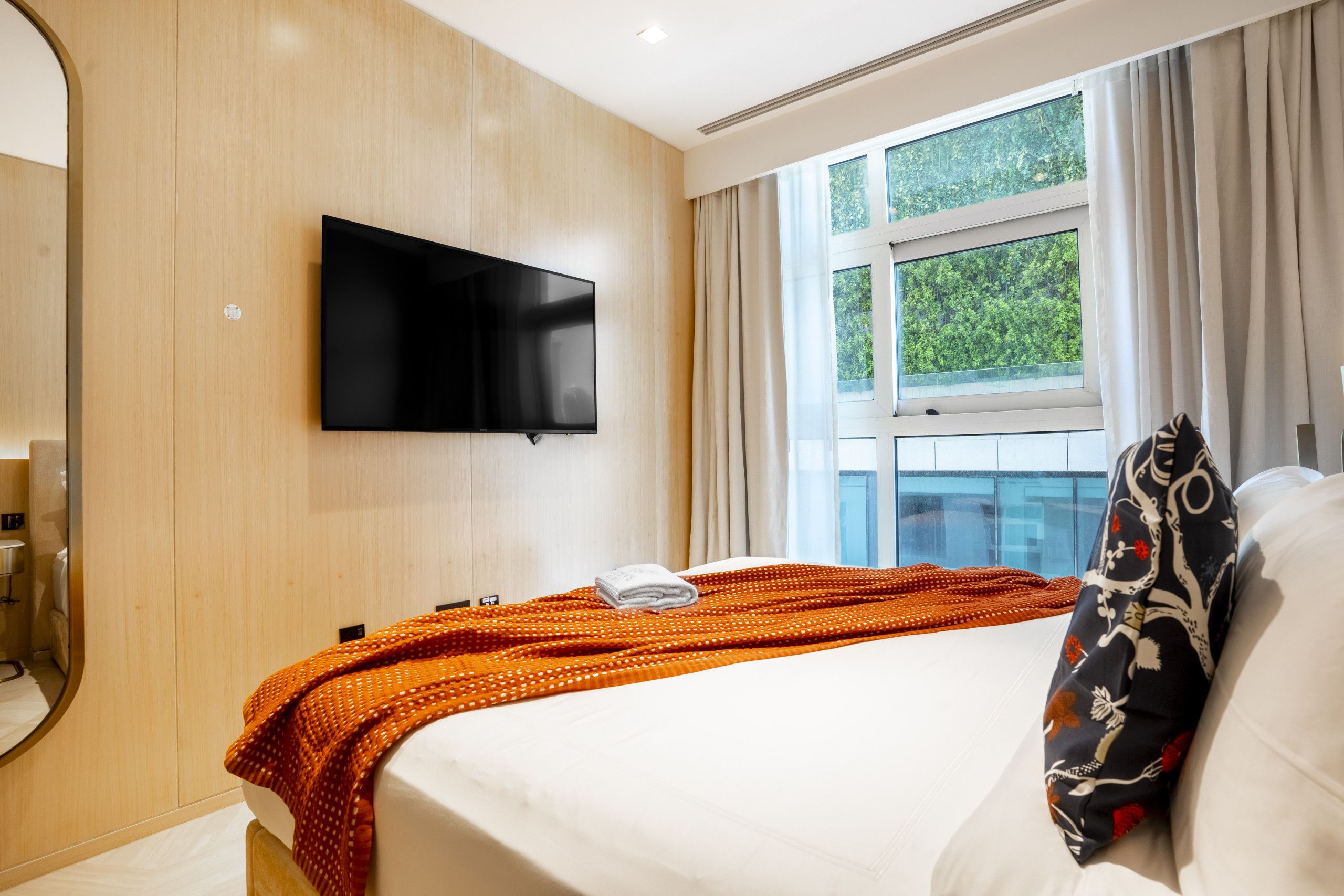 Ett hotellrum med en säng, en platt-TV som är monterad på väggen, ett fönster med gardiner och utsikt över grönområden utanför.