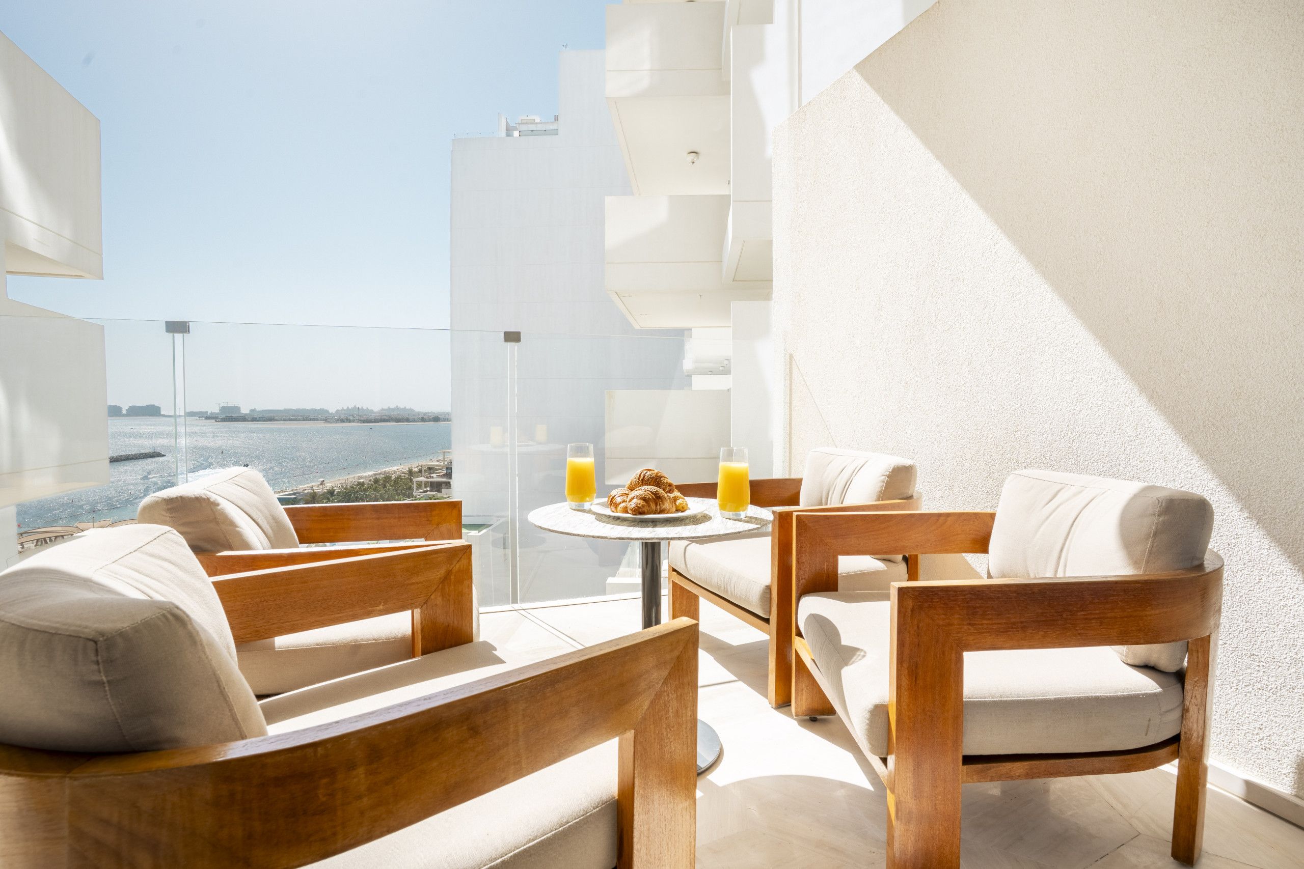 A balcony with a view of the sea, furnished with wooden chairs and a table, and two glasses of orange juice.