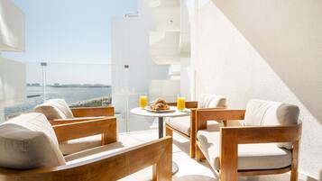 A balcony with a view of the sea, furnished with wooden chairs and a table, and two glasses of orange juice.