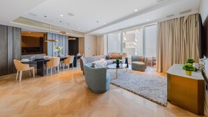 Ein modernes Wohnzimmer mit einer Couch, Sesseln, einem Couchtisch und einem Fernseher. Ein Essbereich mit Stühlen und einem Tisch ist vorhanden, und im Hintergrund befindet sich eine Küche.