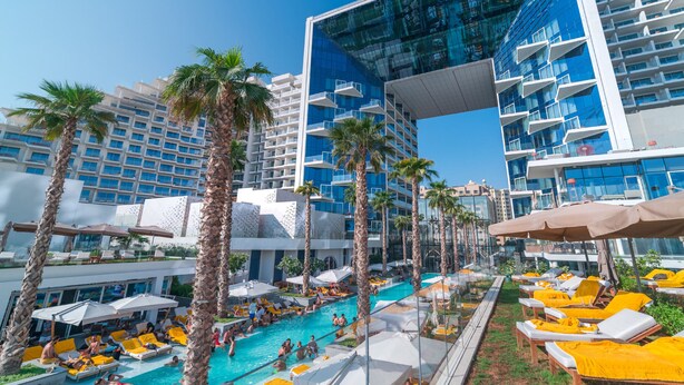 Ein Hotelpoolbereich mit mehreren Balkonebenen, Palmen und Liegestühlen.