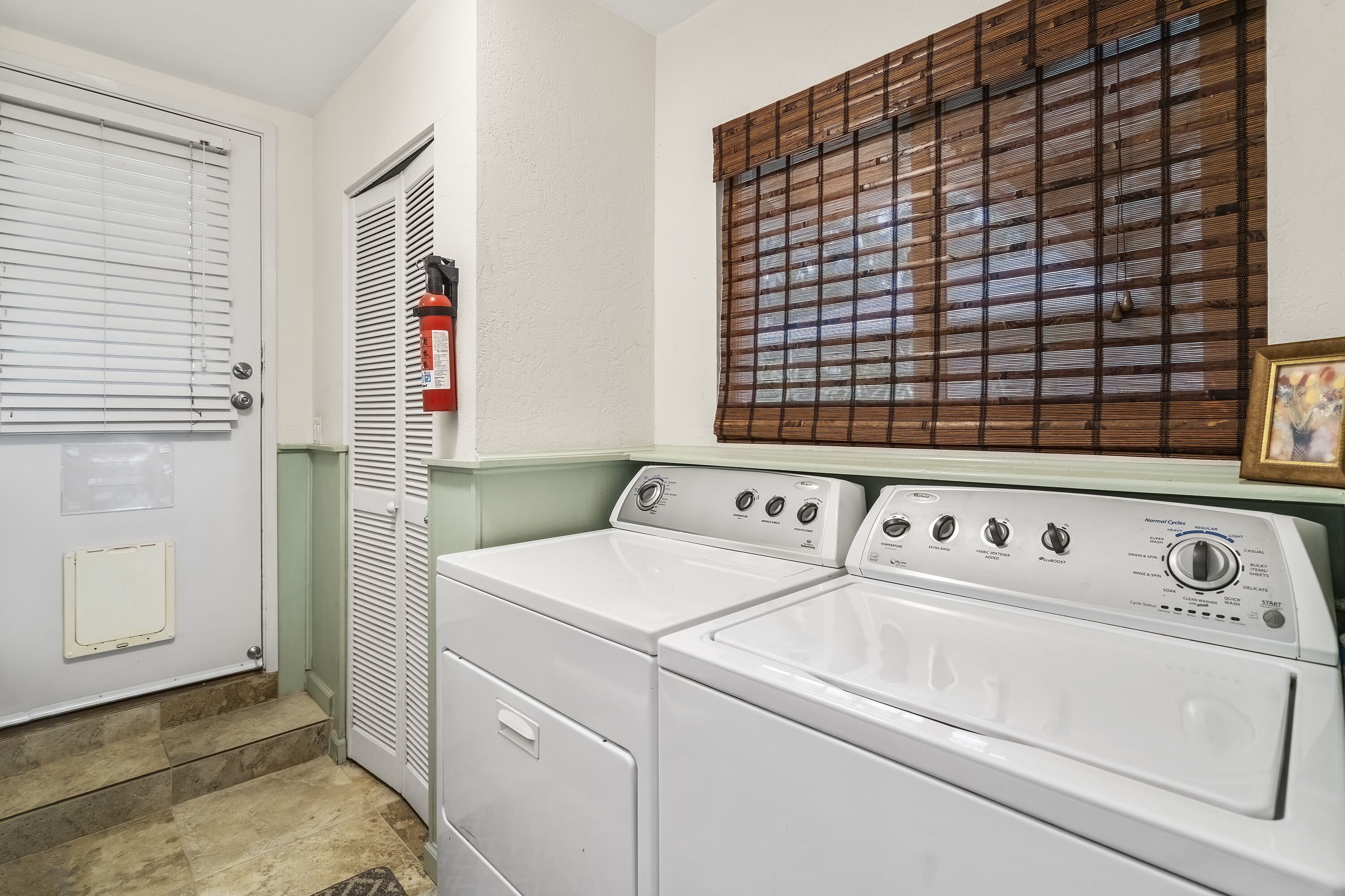 A laundry room with a front-loading washer and dryer, a fire extinguisher, and a window with blinds.