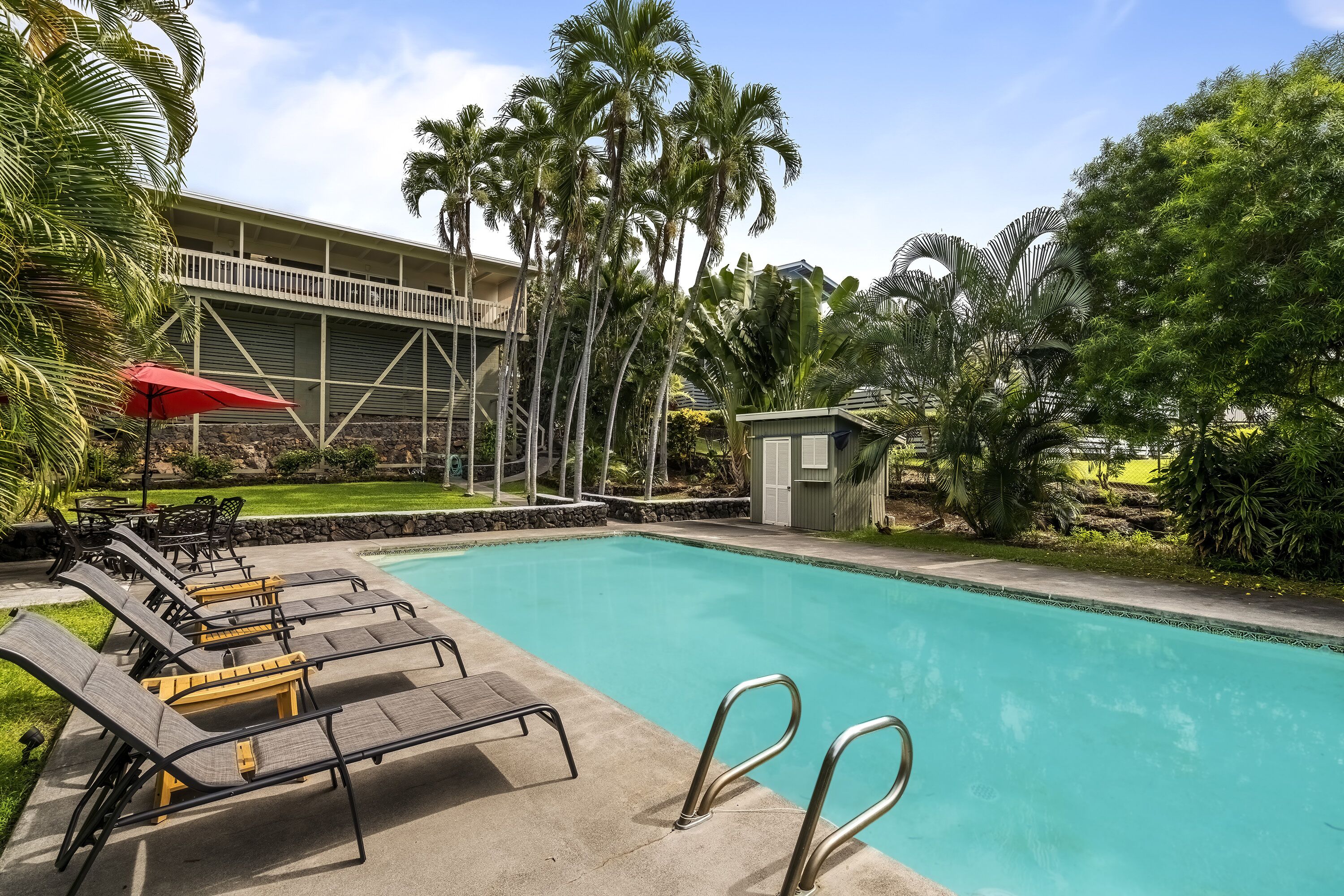 A swimming pool with lounge chairs and a red umbrella, surrounded by palm trees and a building with a balcony.