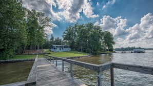 A wooden pier leading to a house by a river, surrounded by trees and a clear sky.