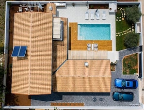 Vista aérea de una propiedad con piscina, tumbonas, un porche cubierto y dos coches aparcados.