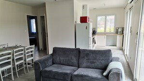 Ein modernes Wohnzimmer mit grauem Sofa, Esstisch und Stühlen. Im Hintergrund ist eine Küche mit Kühlschrank, Mikrowelle und Schränken zu sehen.